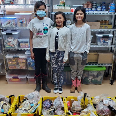 Three children standing in a food pantry surrounded by shelves and baskets full of food.