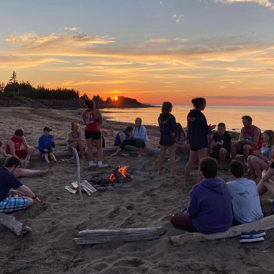 A group of people sitting on the beach around a bonfire.