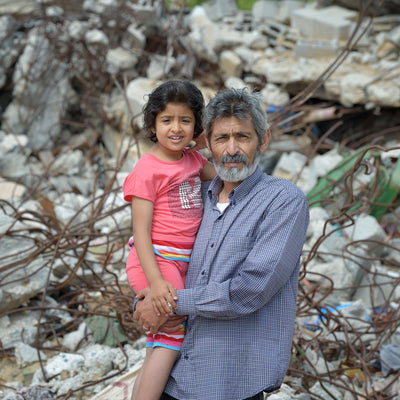 A person holding a child in front of some rubble.